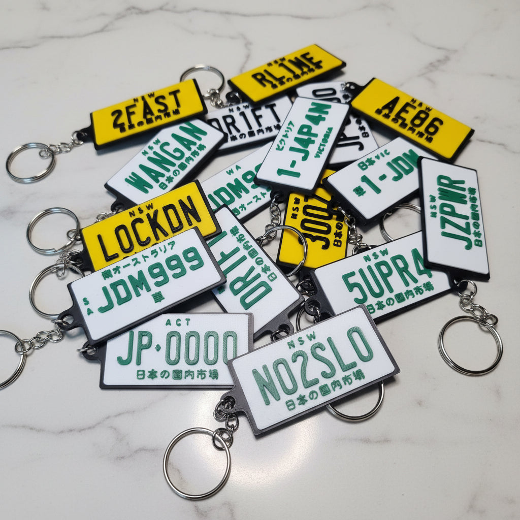 A pile of novelty license plate keychains with silver metal rings, displayed on a marble surface. The keychains feature white plates with green text and yellow plates with green text.