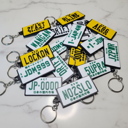 A pile of novelty license plate keychains with silver metal rings, displayed on a marble surface. The keychains feature white plates with green text and yellow plates with green text.
