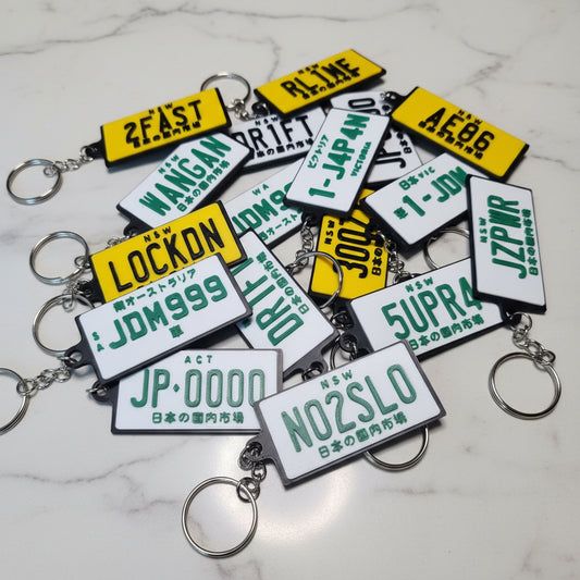 A pile of novelty license plate keychains with silver metal rings, displayed on a marble surface. The keychains feature white plates with green text and yellow plates with green text.