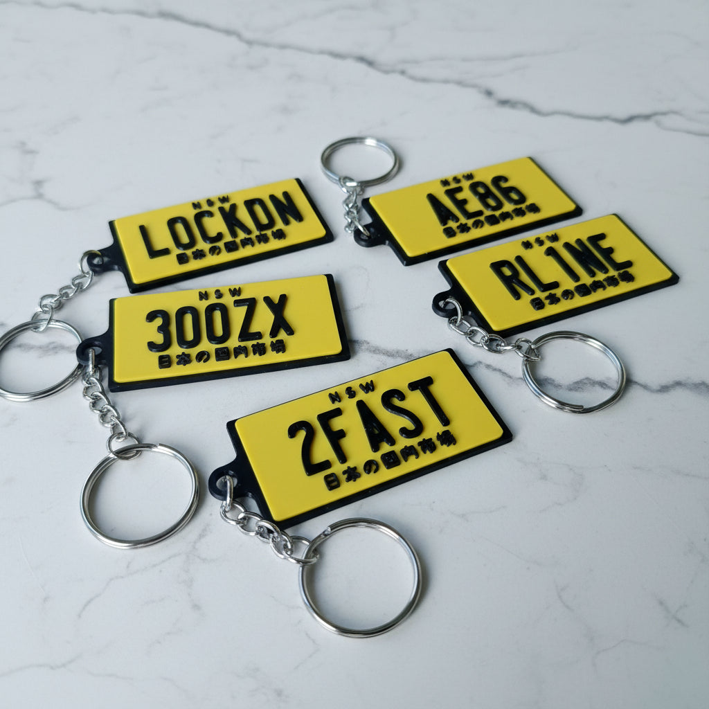 Five yellow novelty license plate keychains with black lettering and silver metal key rings, arranged on a marble surface.