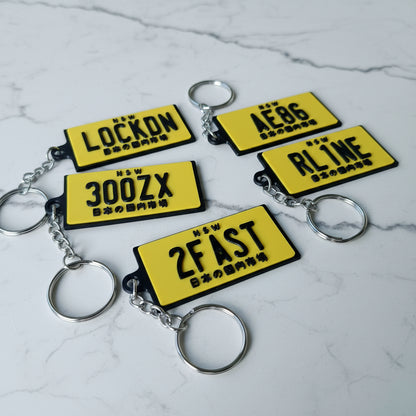 Five yellow novelty license plate keychains with black lettering and silver metal key rings, arranged on a marble surface.