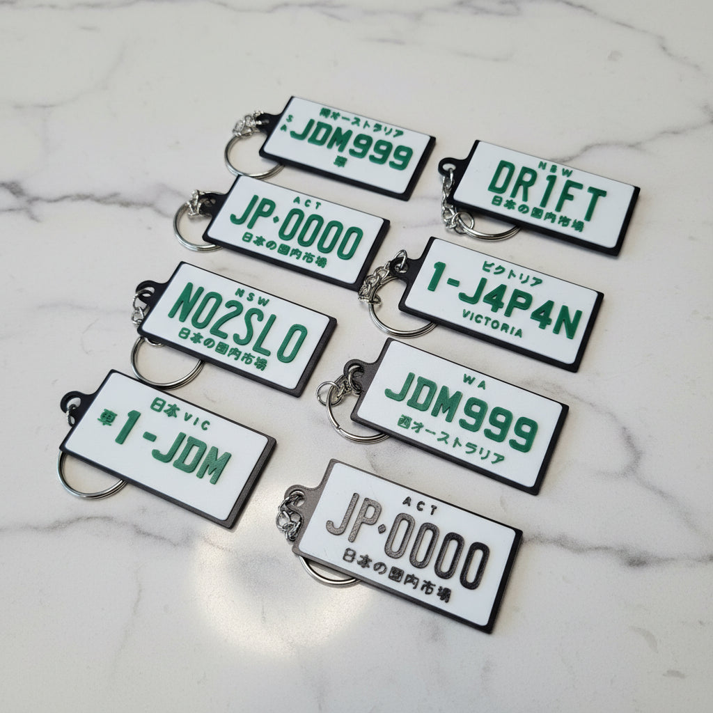 Eight novelty license plate keychains with Japanese designs are arranged on a marble surface. The keychains feature white plates with green and black text, including 'DR1FT', 'JDM999', 'JP-0000', 'N02SL0', '1-J4P4N' and '1-JDM'.