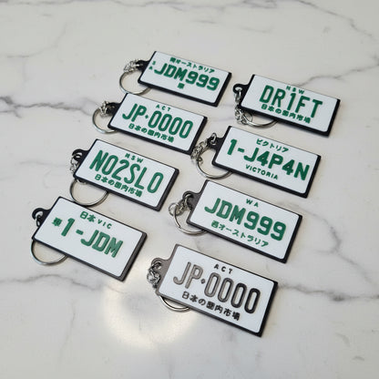 Eight novelty license plate keychains with Japanese designs are arranged on a marble surface. The keychains feature white plates with green and black text, including 'DR1FT', 'JDM999', 'JP-0000', 'N02SL0', '1-J4P4N' and '1-JDM'.
