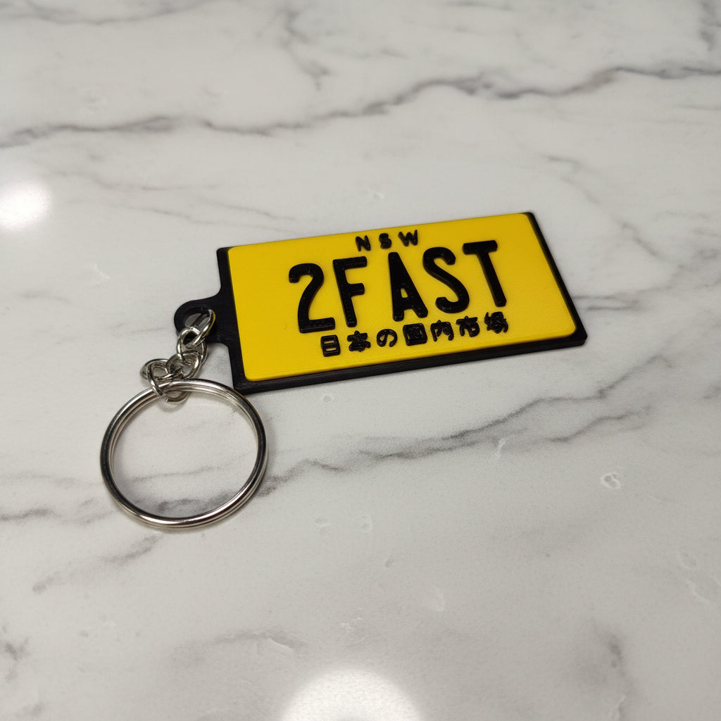 A yellow rectangular novelty keychain with black text reading 'NSW 2FAST' and Japanese characters, attached to a silver metal split ring, shown on a marble background.