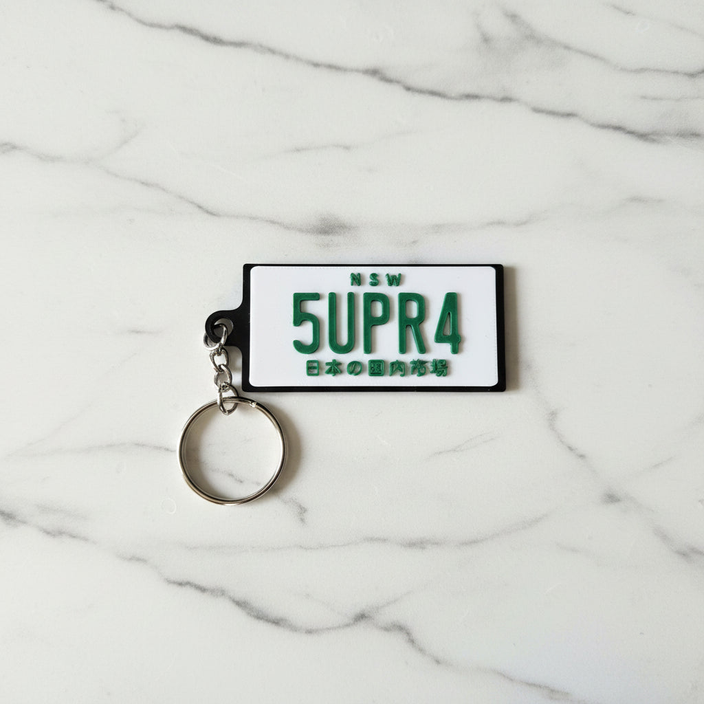 A novelty keychain featuring a white rectangular plate design with green text reading 'NSW 5UPR4' and Japanese characters, attached to a silver metal split ring, set against a marble background.