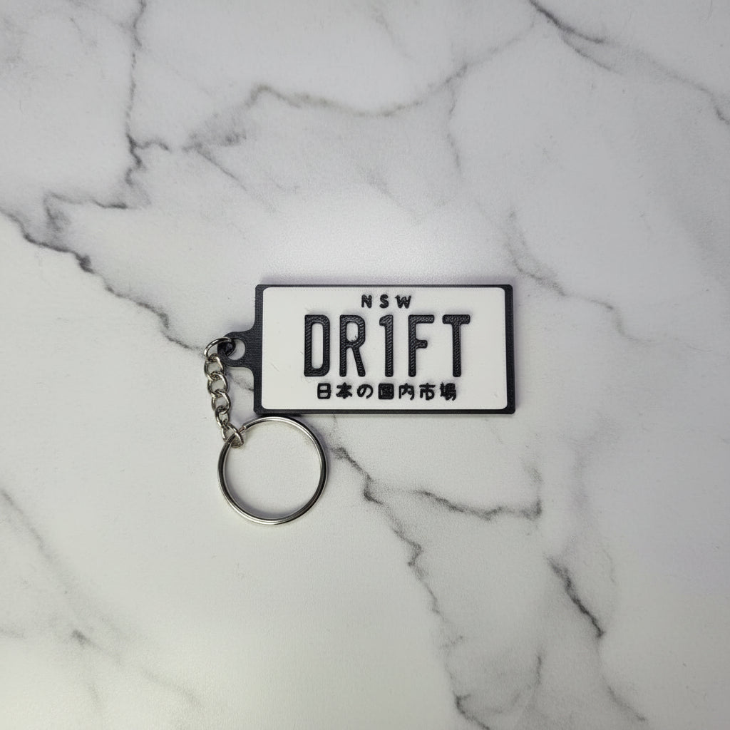 A rectangular white plastic keychain with black text reading 'NSW DRIFT' and Japanese characters, attached to a silver metal key ring on a marble background.