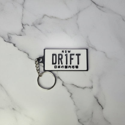 A rectangular white plastic keychain with black text reading 'NSW DRIFT' and Japanese characters, attached to a silver metal key ring on a marble background.