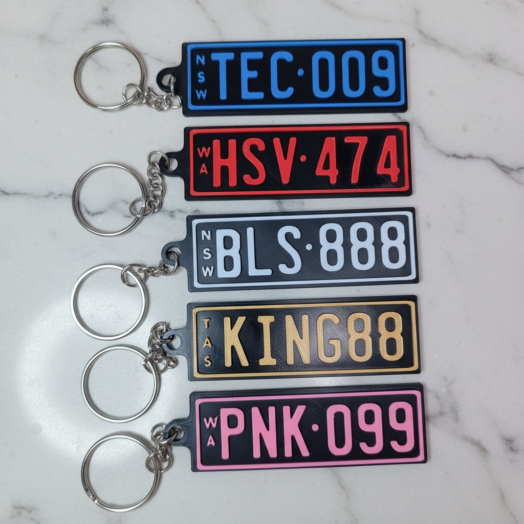 Five rectangular license plate keychains in different colors are arranged in a row on a marble background. The keychains are blue, red, gray, gold, and pink, each with a silver split key ring attached below.