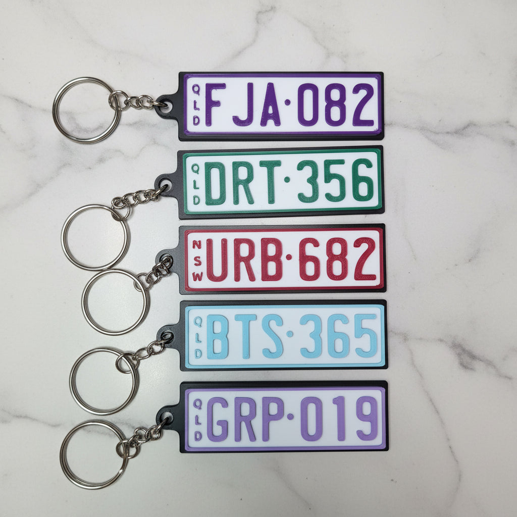 Five rectangular license plate keychains with metal key rings, shown on a marble background. Each plate has a different color and mock registration text.