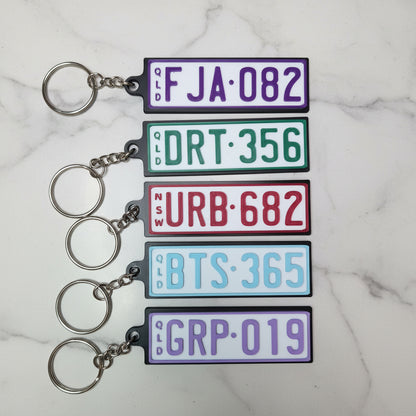 Five rectangular license plate keychains with metal key rings, shown on a marble background. Each plate has a different color and mock registration text.