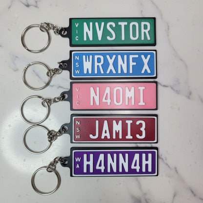 Five rectangular license plate keychains in different colors are lined up on a marble background. The keychains are green, blue, pink, burgundy, and purple, each with white lettering and a silver metal keyring.