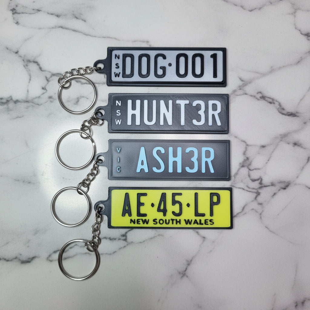 Four novelty license plate keychains in black, gray, and yellow, featuring Australian style plate designs, displayed on a marble background.