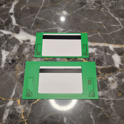 Two green rectangular plastic trim pieces or cover plates with cutouts, displayed against a marble background.