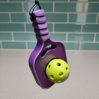 A purple pickleball paddle fidget toy with a shiny handle and a lime green perforated ball, held against a blurred indoor background.