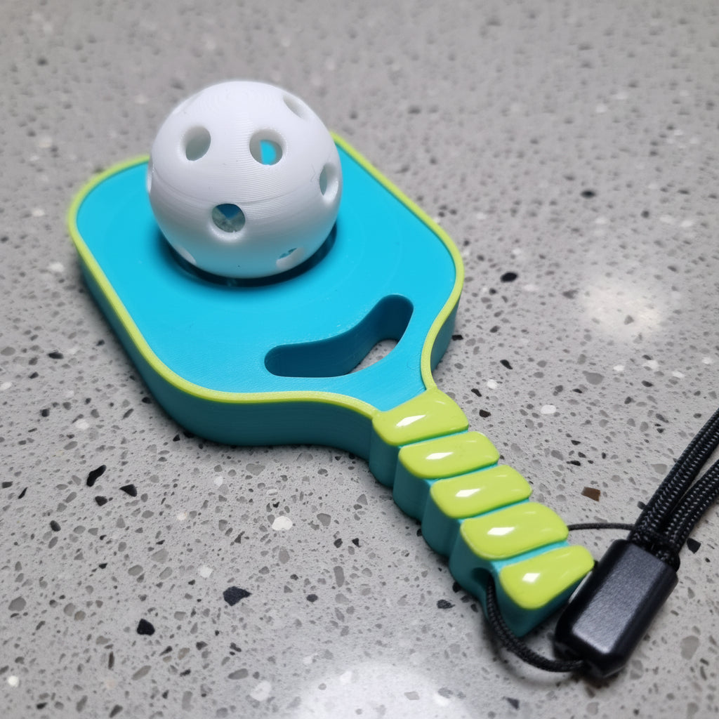 A turquoise coloured pickleball fidget clicker with a lime green edge, a white perforated ball attached, and a lime green handle with a black wrist strap, placed on a grey caesar stone surface.