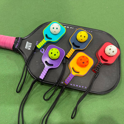 A black pickleball paddlewith a pink handle grip, shown on a green pickleball court floor. Attached to the paddle are five colorful plastic paddle-shaped accessories in purple, blue, yellow, navy, and red, each with a black wrist strap and a small round colored button.